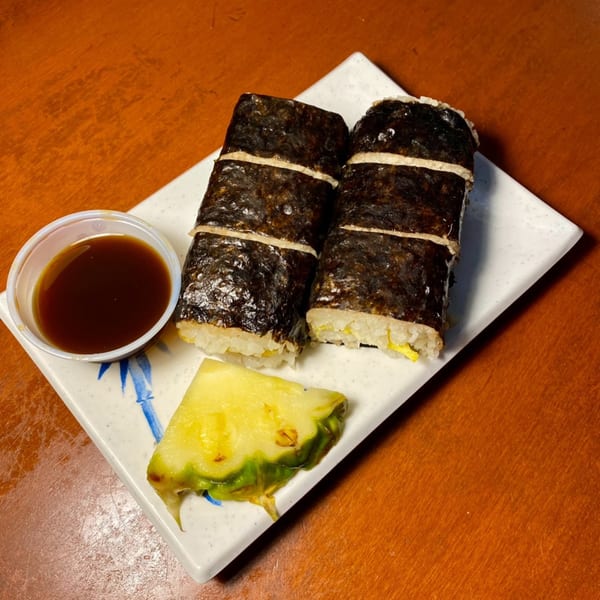 Two Hawaiian musubi rolls with a side of soy sauce and a fresh pineapple slice.