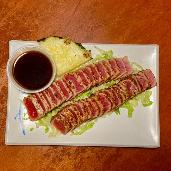 Seared tuna slices on lettuce with a side of dark sauce and a pineapple wedge.